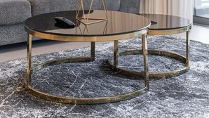 Ein modernes Wohnzimmer mit einem grauen Sofa, einem runden Glastisch mit goldenen Beinen und einer hohen, grünen Pflanze im schwarzen Topf.