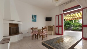 A room with a fireplace, a dining table with chairs, a TV mounted on the wall, and a large window with a view of greenery. - 3599 Casa Delma by Barbarhouse (Porto Cesareo)