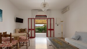 A modern living room with a sofa, a coffee table, a dining table with chairs, a TV, and a view through an open doorway to a garden. - 3599 Casa Delma by Barbarhouse (Porto Cesareo)