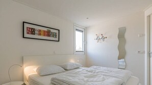 Ein schlicht eingerichtetes Schlafzimmer mit einem ordentlich bezogenen Bett, einem Nachttisch mit Lampe, einem Fenster und einer Wandleuchte.