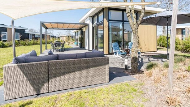 Ein moderner Außenbereich mit einer großen Rattan-Couch, einer überdachten Terrasse und einem gläsernen Gebäude im Hintergrund.