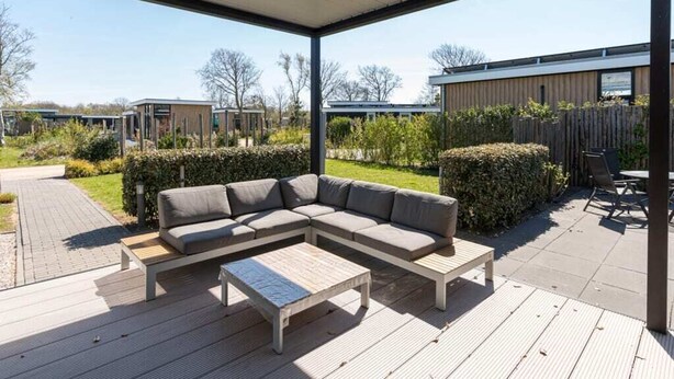 An outdoor patio with a sectional sofa and a coffee table, surrounded by a wooden deck and a covered area. - Discover this beautiful L-shaped chalet with a covered, sheltered terrace and a lovely lounge set. T (Burgh-Haamstede)