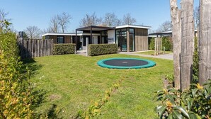 A modern house with a well-maintained lawn, a trampoline, and a wooden fence. - Discover this beautiful L-shaped chalet with a covered, sheltered terrace and a lovely lounge set. T (Burgh-Haamstede)