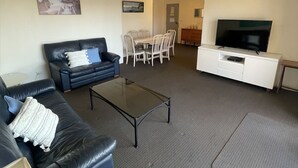 Un salon avec un canapé en cuir noir, une table basse en verre, un meuble TV blanc et un coin repas avec des chaises et une table blanches.
