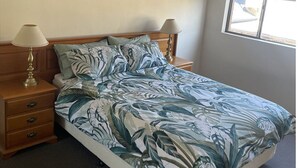 Un lit bien fait, recouvert d’une couvre-lit à motifs tropicaux, avec deux lampes de chevet et une table de chevet en bois dotée de tiroirs.