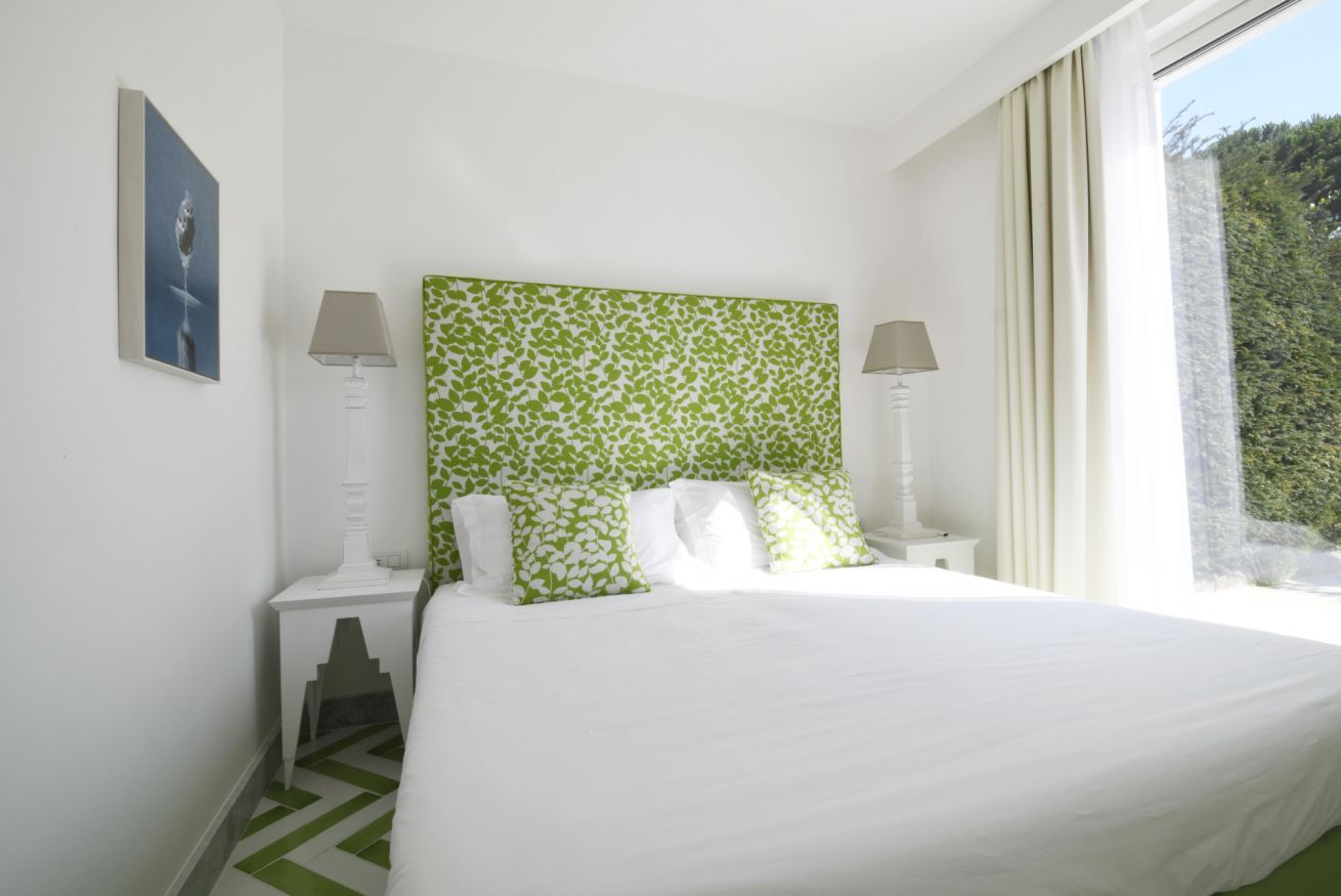 Une chambre d’hôtel avec un lit, deux tables de chevet, une tête de lit à motifs verts et une fenêtre avec des rideaux.