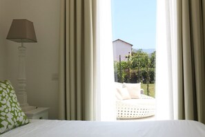 Une chambre à coucher avec un lit, une table de chevet, une lampe et une vue sur un jardin et un bâtiment, le tout visible à travers des rideaux transparents.