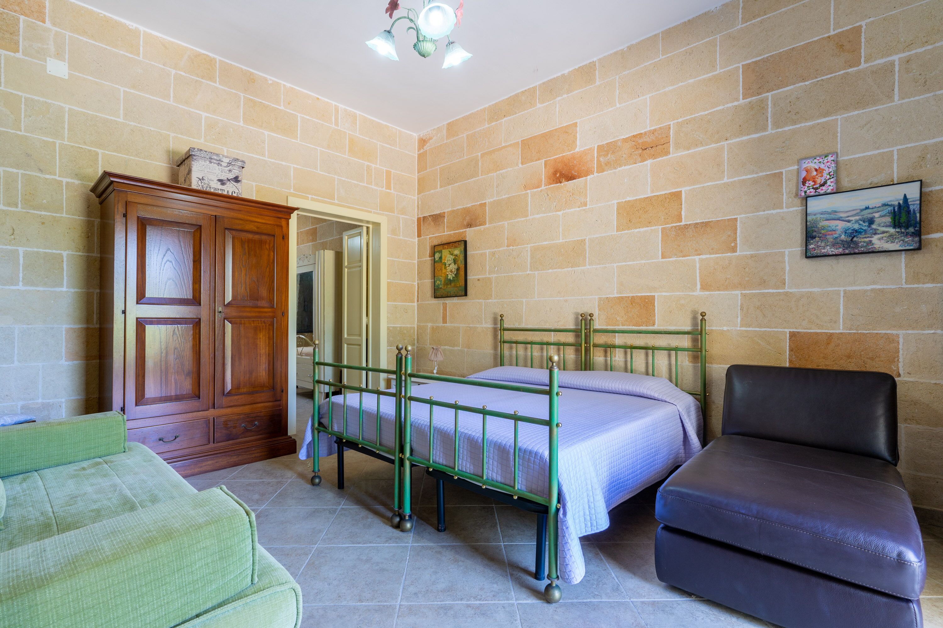A room with a bed, a green metal frame, a wooden wardrobe, a green sofa, and a black leather chair.