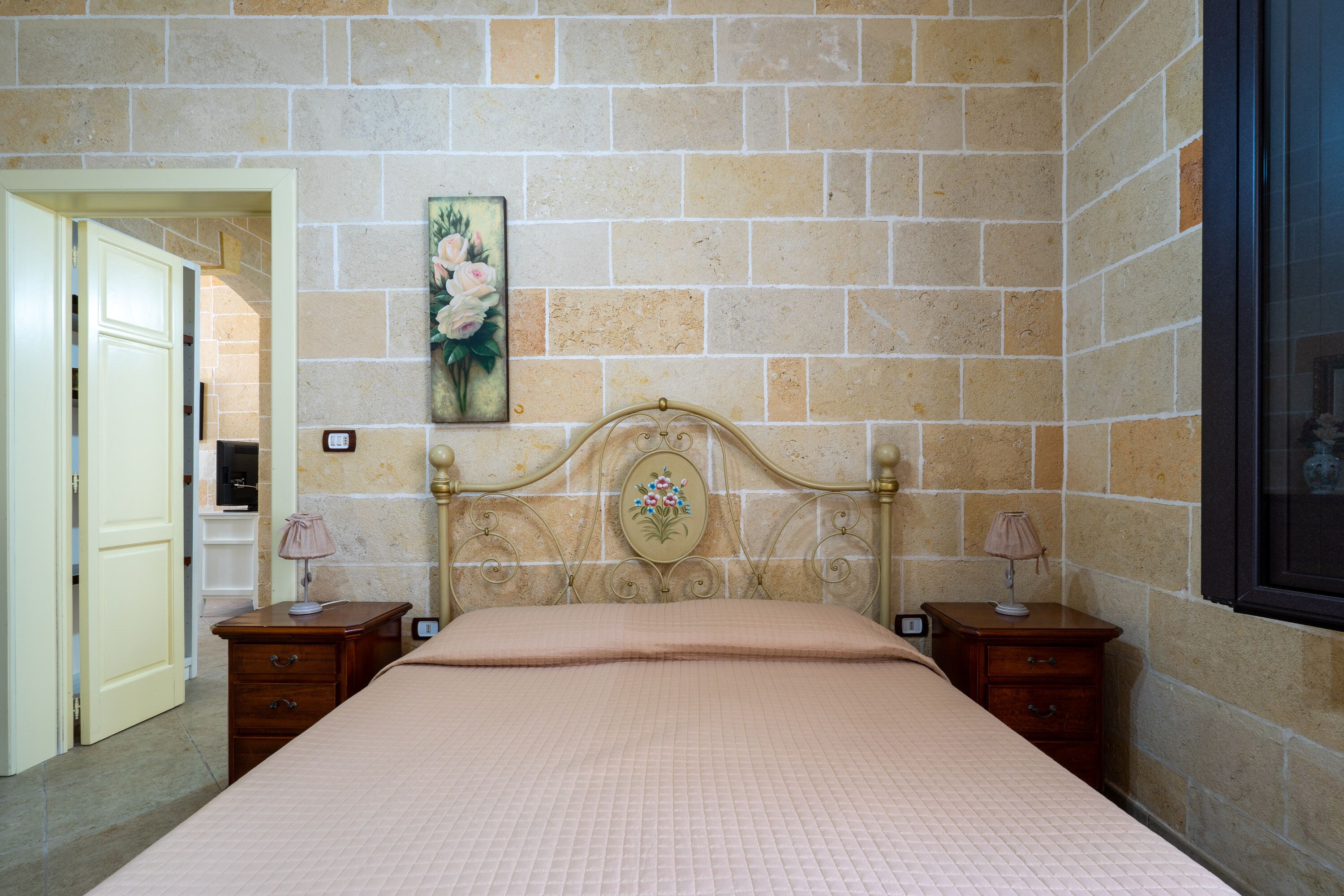 A bedroom with a bed, two nightstands, a painting on the wall, and a door leading to another room.