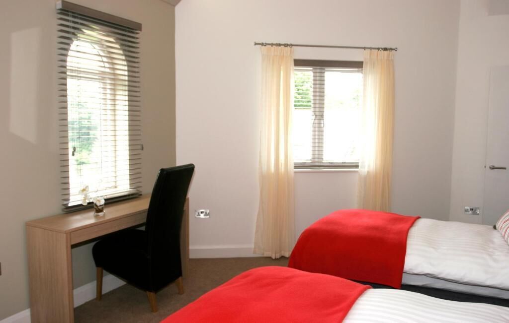 Une chambre avec un bureau, une chaise, un lit avec une literie rouge et blanche, et une fenêtre avec des rideaux.