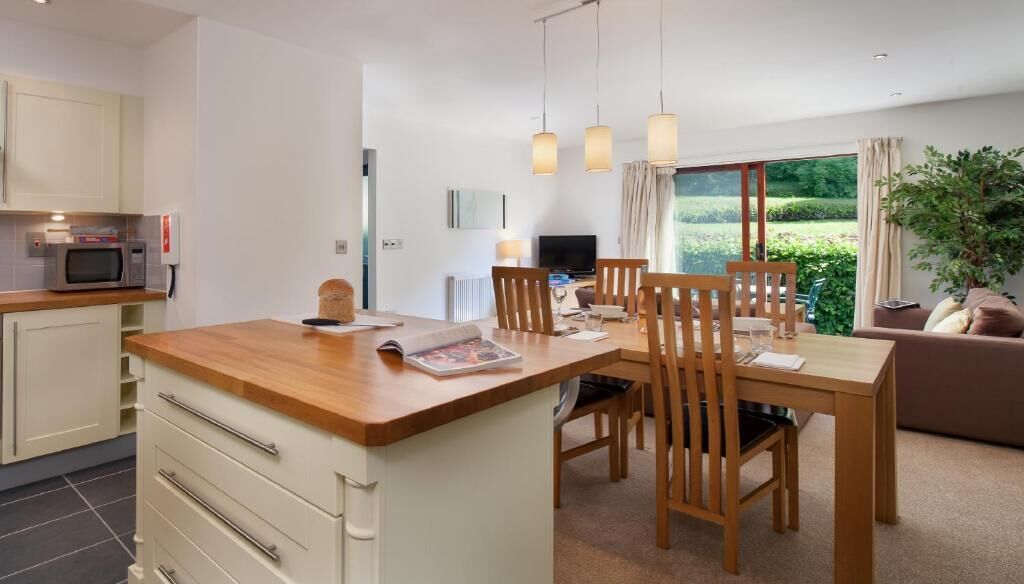A modern kitchen with a wooden island, dining area with wooden chairs, a TV, and a large window with a view of greenery.