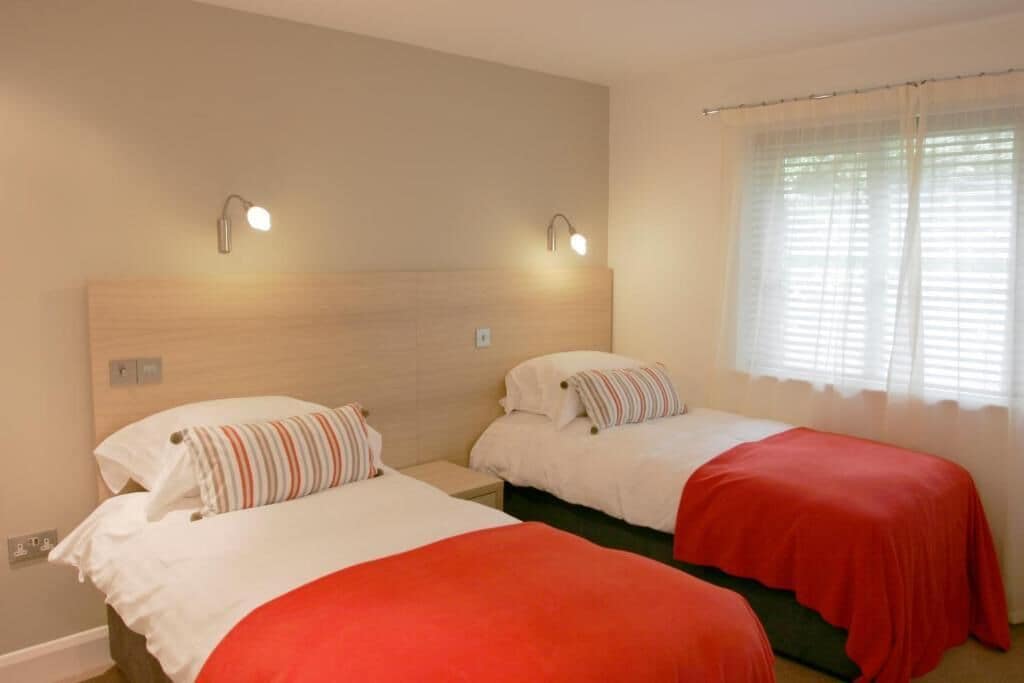 A hotel room with two single beds, each with a red bedspread and striped pillows. There are wall-mounted lights above each bed and a window with sheer curtains.