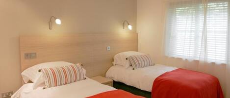 A hotel room with two single beds, each with a red bedspread and striped pillows. There are wall-mounted lights above each bed and a window with sheer curtains.