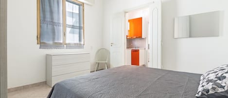 A bedroom with a bed, a window with curtains, a white dresser, and an open door leading to a kitchen with orange cabinets.