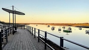 Een houten steiger met leuningen, banken en parasols, met uitzicht op kalm water en boten.