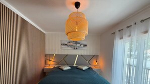 Een slaapkamer met een groot bed, twee bedlampen, een hanglamp en een raam met dunne gordijnen.
