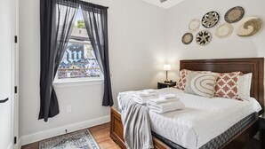 Una cama bien hecha con ropa blanca, un cabecero de madera y una mesita de noche con lámpara. Se ve una ventana con cortinas negras y vistas a edificios.
