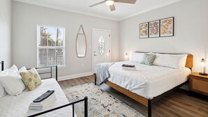 Un dormitorio con cama, mesitas de noche, ventilador de techo, ventana con persianas, puerta con espejo decorativo y decoración en la pared.
