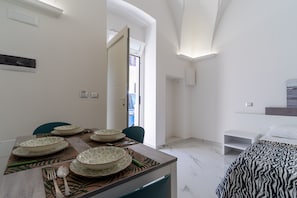 Et moderne hotelværelse med et spiseområde, der indeholder et træbord, mønstrede underlag og matchende skåle. En seng med et sengetæppe med zebraprint er synlig, og værelset har hvide vægge og et marmorgulv.
