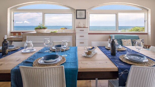A dining area with a view of the sea, featuring a wooden table set with plates, glasses, and a decorative plant.