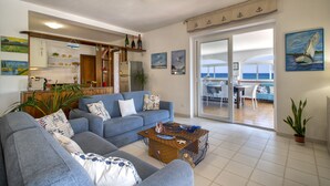 A living room with a sofa, a coffee table, and a view of the sea through a sliding glass door.