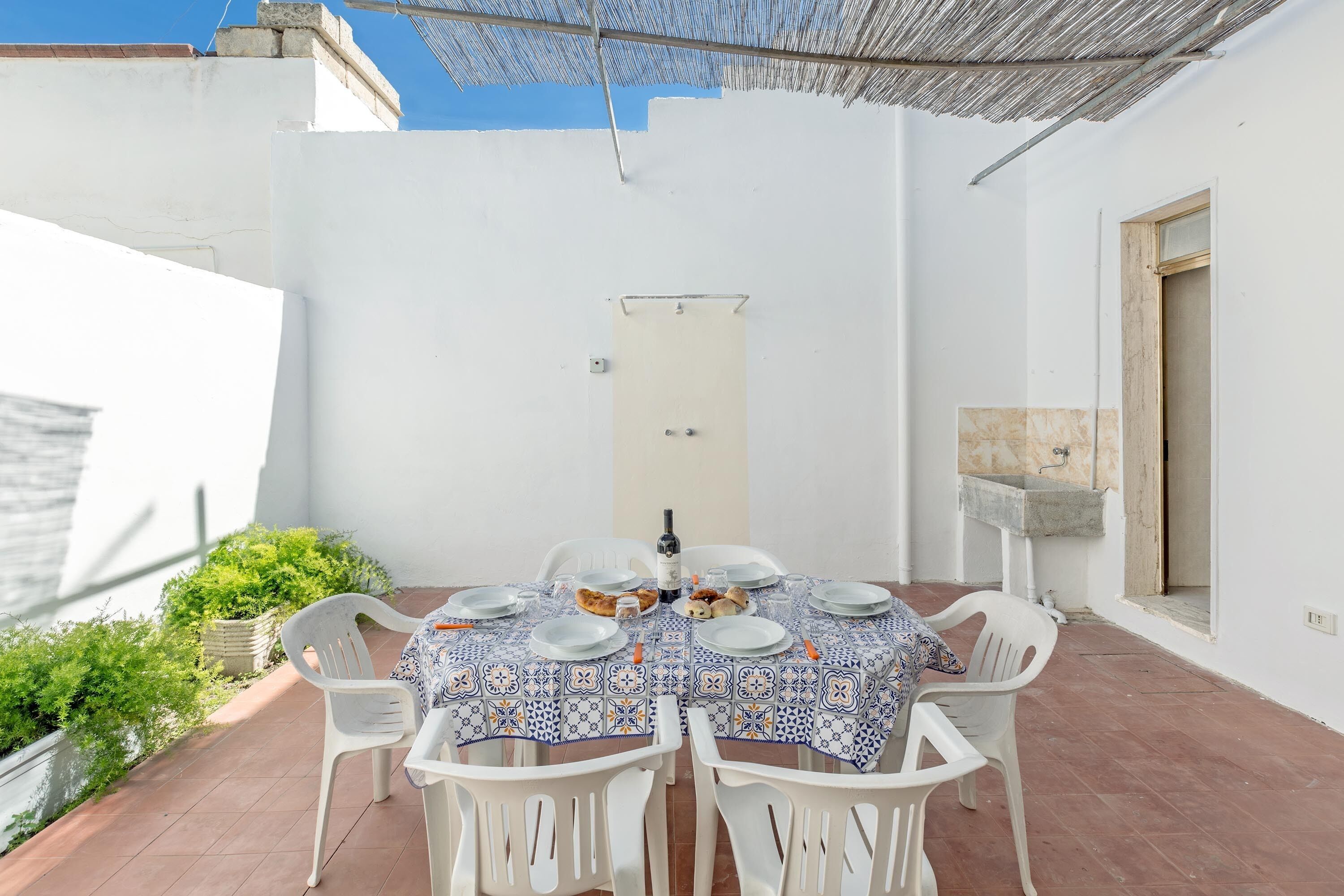 A patio with a table set for two, white plastic chairs, and a tiled tablecloth.