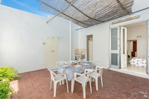 Un patio con una mesa lista para cuatro personas, rodeado de sillas blancas, bajo una zona sombreada.