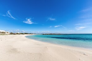 Ein weiter Sandstrand mit klarem türkisblauem Wasser und ein paar Gebäuden in der Ferne.