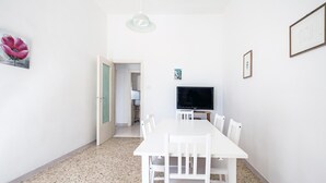 Un comedor con una mesa y sillas blancas, un televisor sobre un mueble bajo y una puerta que da a otra habitación.