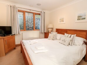 A neatly made bed with white linens and pillows, a television on a wooden cabinet, a window with curtains, and a lamp on a bedside table. - 1 Court Cottage, Hillfield Village (Dartmouth)