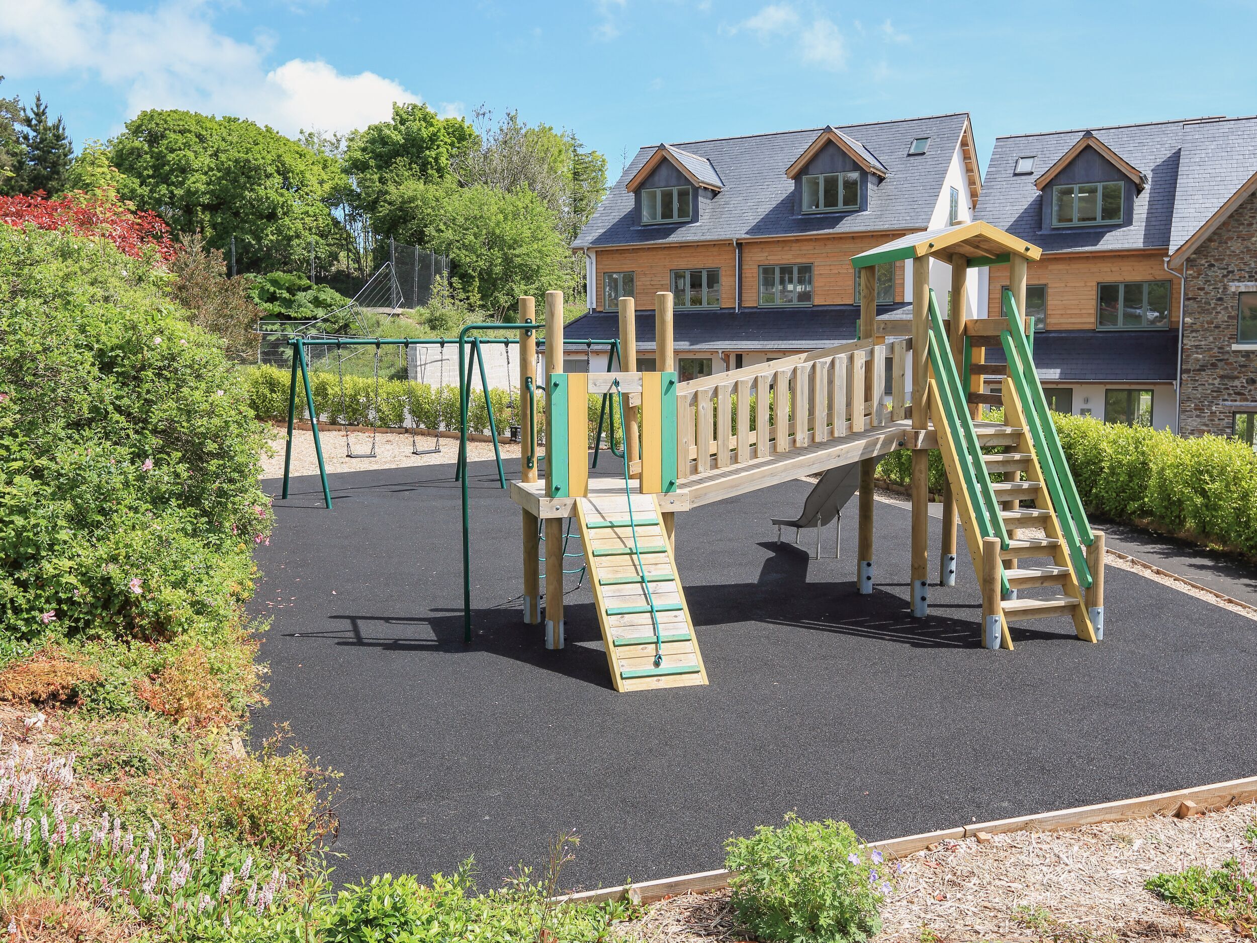 A playground with wooden equipment, surrounded by a landscaped garden and a building with a sloped roof.