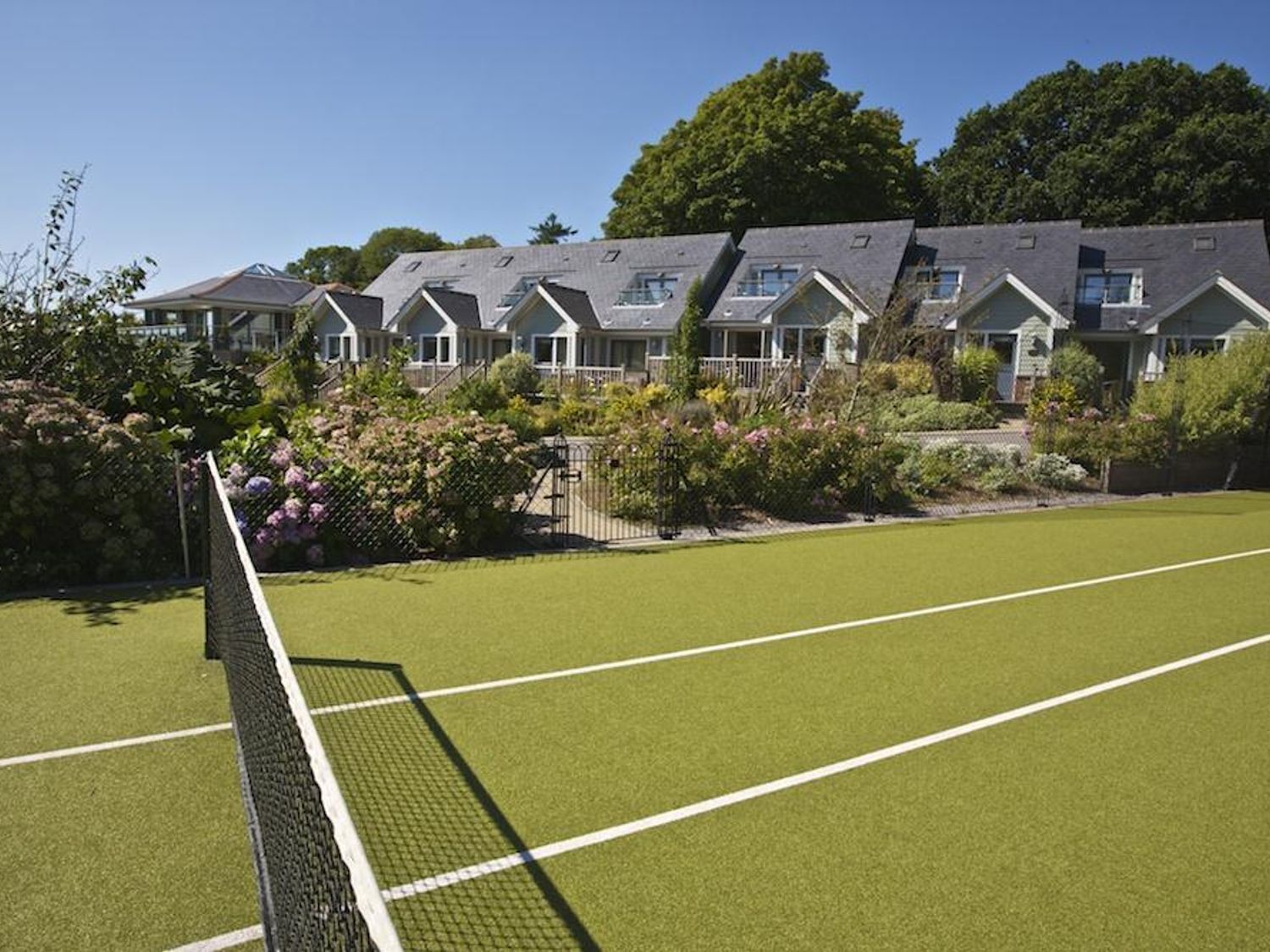 Una cancha de tenis con red, rodeada por una hilera de casas con techos inclinados y un jardín cuidado.