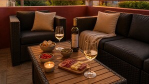 Un balcón con muebles de mimbre, una botella de vino, copas y una tabla de embutidos con aceitunas, queso y carnes.