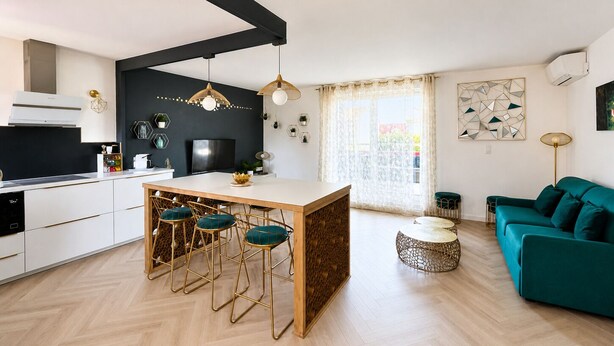 Una cocina moderna con una isla central, taburetes de bar, una mesa de comedor, un sofá y un elemento decorativo en la pared.