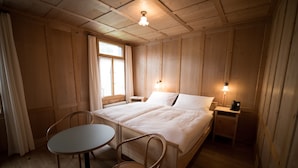 A wooden room with a bed, a table, two chairs, a window with curtains, a bedside lamp, and a radiator.
