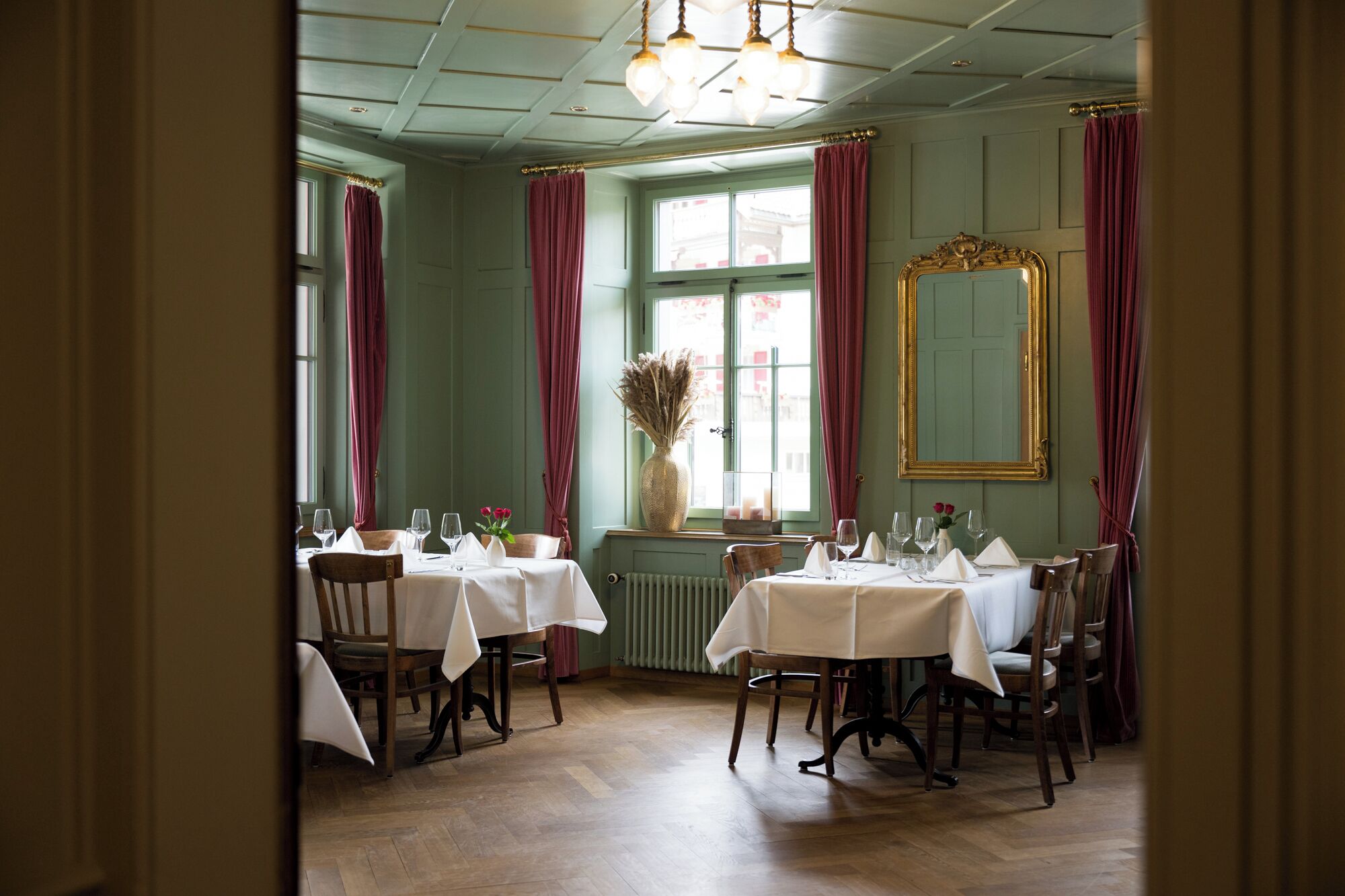 Un comedor con mesas dispuestas para una comida, un espejo grande y una ventana con cortinas.