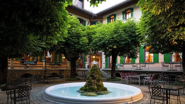 A courtyard with a central fountain, surrounded by outdoor seating and a building with green shutters in the background.