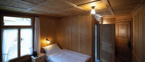 Ein Zimmer mit Holzboden, einem Bett, einem Nachttisch, einem Fenster mit Vorhängen und einer Tür.