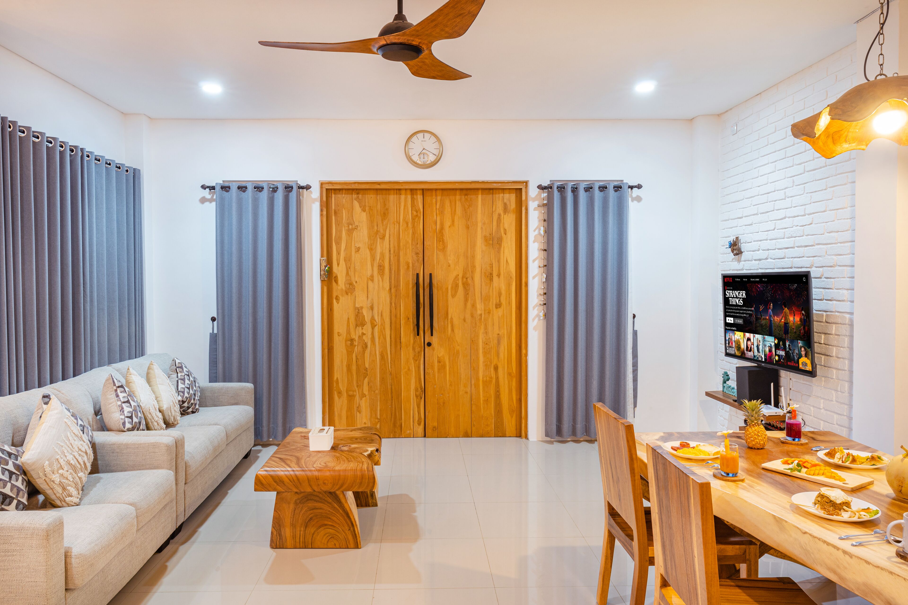 Uma sala de estar moderna com um sofá, mesa de centro, mesa de jantar e cadeiras. Há uma porta de madeira com cortinas, um ventilador de teto e uma TV na parede.