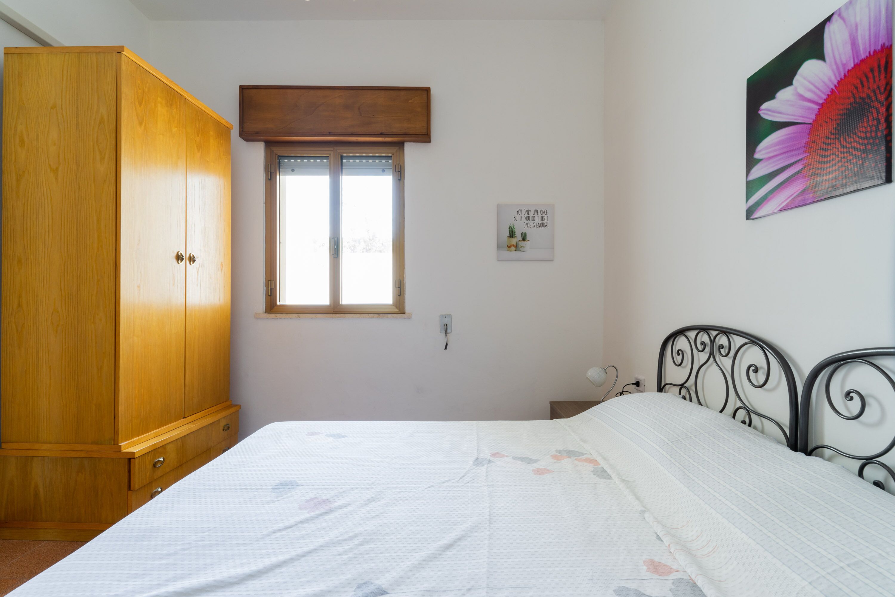 A bedroom with a wooden wardrobe, a bed with a floral bedspread, a window with a wooden blind, and a wall-mounted picture of a flower.