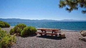 A beach with a wooden picnic table, surrounded by rocks and greenery, overlooking the sea and distant mountains.