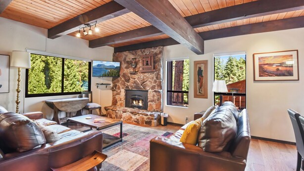 A cozy living room with a stone fireplace, leather sofas, and large windows offering views of greenery.