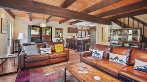 A spacious living area with leather sofas, a coffee table, and a rug. There is a kitchen in the background with a dining area.
