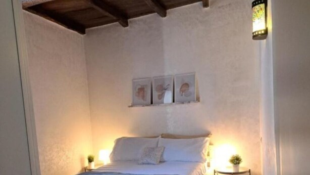 Une chambre avec un plancher en bois, un lit avec une literie blanche, une petite table de chevet avec une lampe et une étagère murale ornée de trois tableaux encadrés.