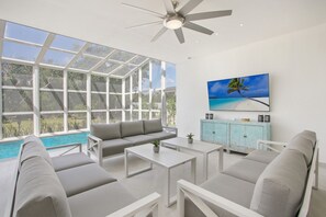 A modern living room with a large window, a ceiling fan, a TV, and a pool view. - Palmas 15: Luxury villa w/ mosquito net, lap-pool, Jacuzzi, staff & golf cart (Punta Cana)