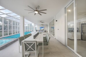 Ein moderner Poolbereich im Innenbereich mit einem langen Tisch und Stühlen, Deckenventilatoren und einem Fernseher.
