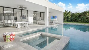 Et moderne hus med swimmingpool, spiseplads og stort vindue med udsigt over grønne områder.