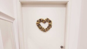 Una puerta blanca con un adorno de guirnalda con forma de corazón y un felpudo con la inscripción 'hereinpazient'.