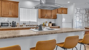 A modern kitchen with wooden cabinets, a stainless steel range hood, a granite countertop, and a bar area with yellow chairs. - Sun & Fun, Backyard and Pool (Tempe)