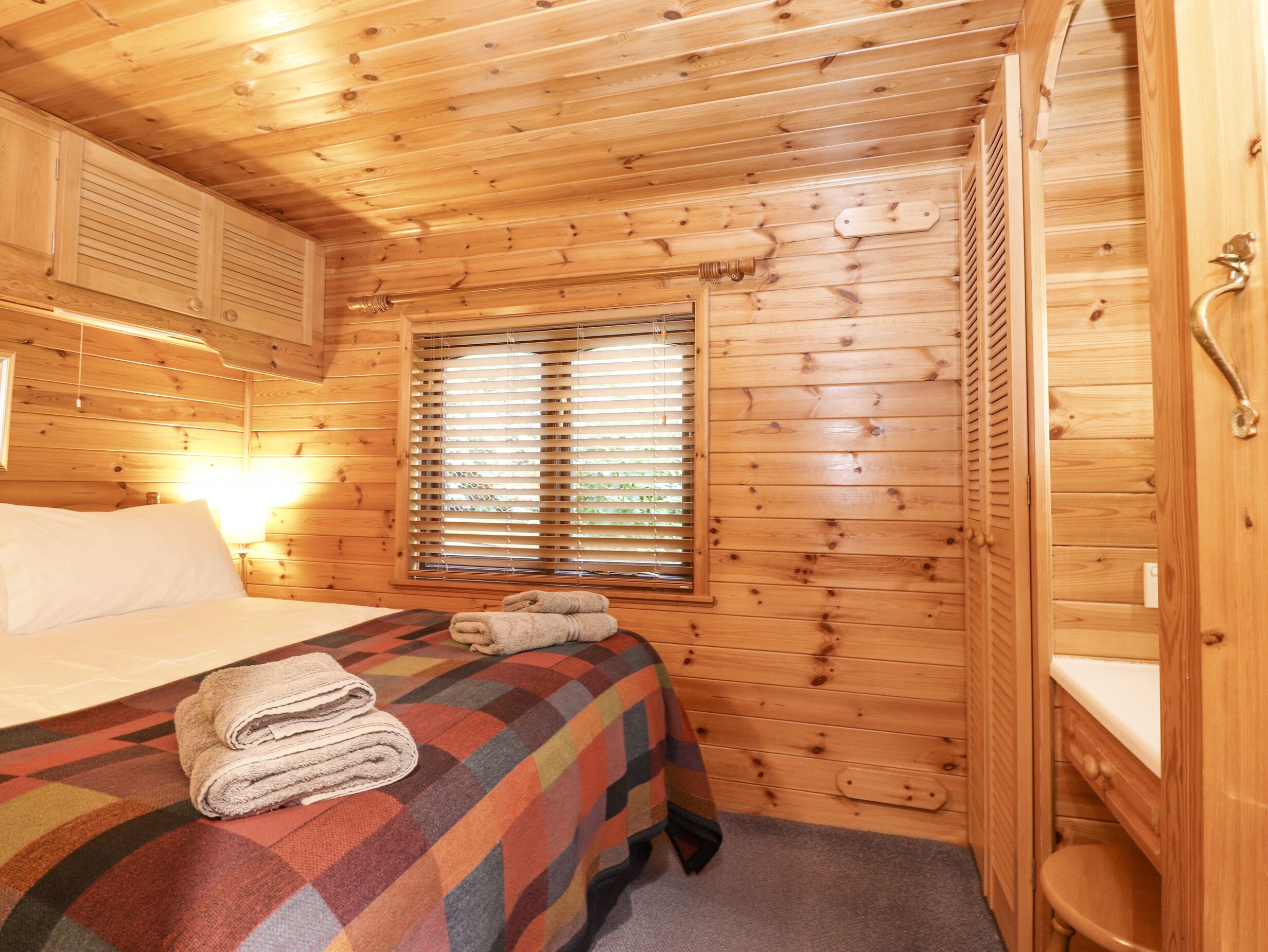 Une pièce de type cabane en bois comprenant un lit, une fenêtre avec des stores, une porte en bois et un petit espace salle de bain.